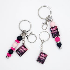 May include: Two keychains with silver rings and lobster clasps. Each keychain features a charm with the text "Butcher & Blackbird" and a beaded accent in pink, black, and silver. The keychains are displayed on a white background.