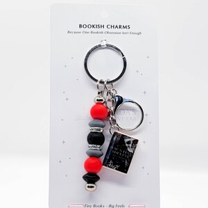 May include: A keychain with a black ring, red, gray, and black beads, and a miniature book charm. The book charm has a black and white cover. The text "BOOKISH CHARMS" and "Tiny Books - Big Feels" are visible on the card.