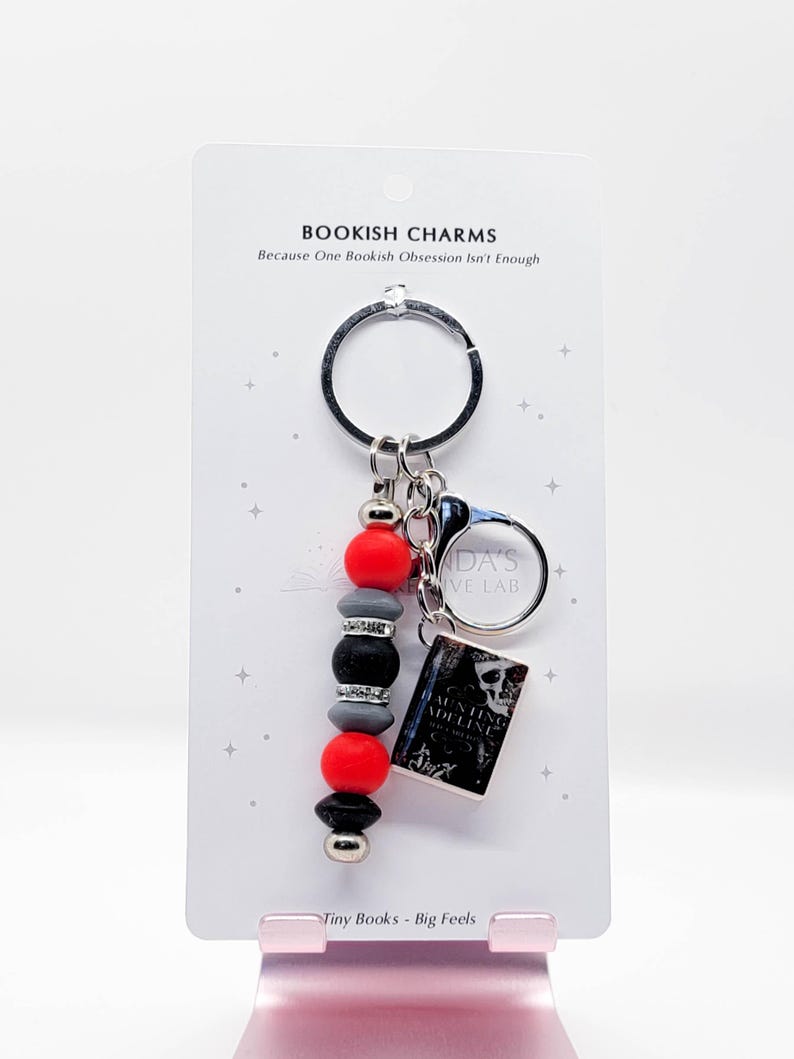 May include: A keychain with a black ring, red, gray, and black beads, and a miniature book charm. The book charm has a black and white cover. The text "BOOKISH CHARMS" and "Tiny Books - Big Feels" are visible on the card.