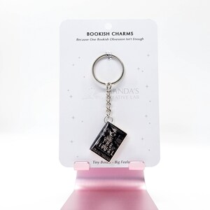 May include: A keychain featuring a miniature book charm with the title "The Ritual" and the author's name. The charm hangs from a silver keyring and chain, displayed on a white card with the text "Bookish Charms". The card is held by a pink stand.