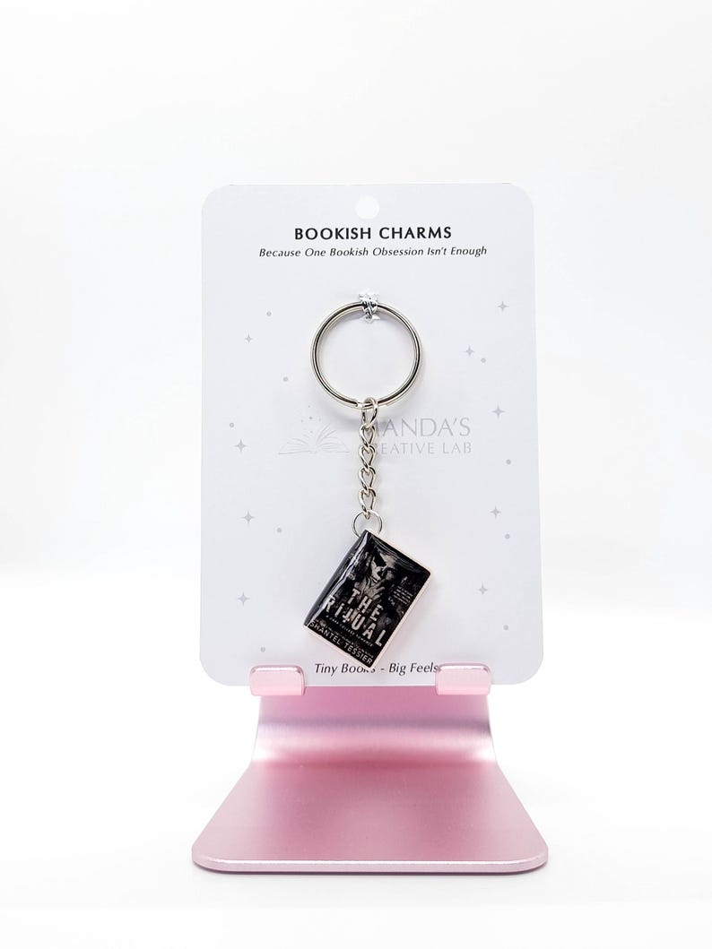 May include: A keychain featuring a miniature book charm with the title "The Ritual" and the author's name. The charm hangs from a silver keyring and chain, displayed on a white card with the text "Bookish Charms". The card is held by a pink stand.
