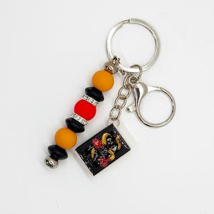 May include: A keychain with a black rectangular charm featuring a gold ram and red roses. The keychain has a beaded strand with orange, red, black, and silver beads. A silver key ring and clasp complete the design.