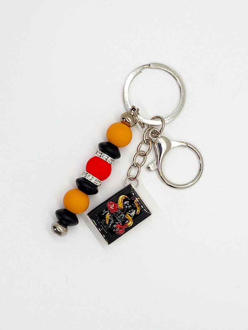 May include: A keychain with a black rectangular charm featuring a gold ram and red roses. The keychain has a beaded strand with orange, red, black, and silver beads. A silver key ring and clasp complete the design.