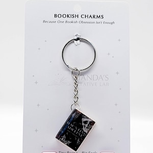 May include: A silver keychain with a miniature book charm. The book cover features a skull and the title "Haunting Adeline". The card behind the keychain reads "Bookish Charms" and "Tiny Books - Big Feels".