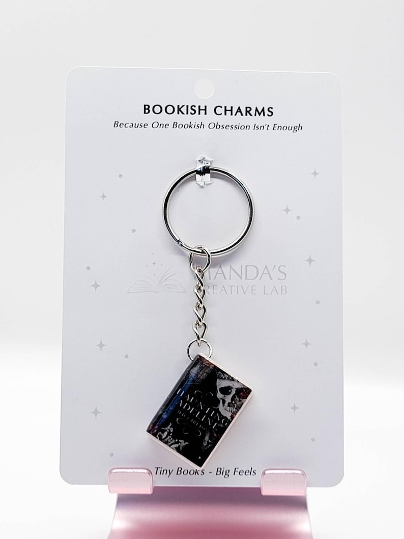 May include: A silver keychain with a miniature book charm. The book cover features a skull and the title "Haunting Adeline". The card behind the keychain reads "Bookish Charms" and "Tiny Books - Big Feels".