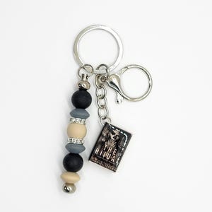 May include: A silver keychain with a beaded accent in black, gray, and beige. The keychain includes a small rectangular charm with the text "THE RITUAL" and "SHANTEL TESSIER". The keychain has a lobster clasp and a key ring.