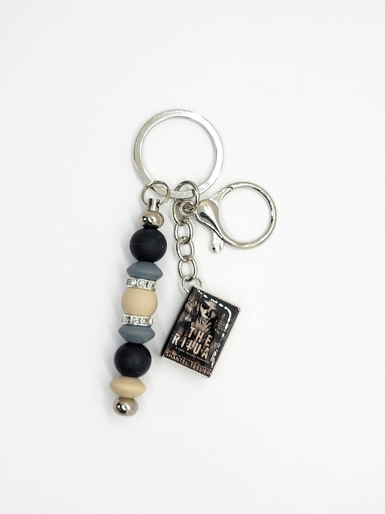 May include: A silver keychain with a beaded accent in black, gray, and beige. The keychain includes a small rectangular charm with the text "THE RITUAL" and "SHANTEL TESSIER". The keychain has a lobster clasp and a key ring.
