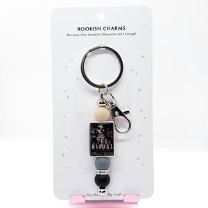 May include: A keychain featuring a small book charm with the title "The Ritual" and a black and white cover image. The keychain includes a silver ring, a lobster clasp, and decorative beads in cream, gray, and black. The card reads "BOOKISH CHARMS Because One Bookish Obsession Isn't Enough."