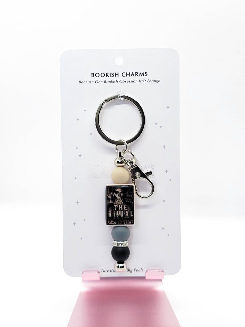 May include: A keychain featuring a small book charm with the title "The Ritual" and a black and white cover image. The keychain includes a silver ring, a lobster clasp, and decorative beads in cream, gray, and black. The card reads "BOOKISH CHARMS Because One Bookish Obsession Isn't Enough."