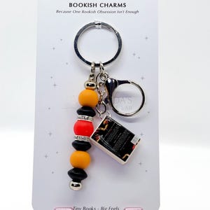 May include: A keychain with a silver ring and a charm featuring a miniature book and a beaded accent. The beads are black, orange, and red, with silver accents. The card reads "BOOKISH CHARMS" and "Tiny Books - Big Feels."