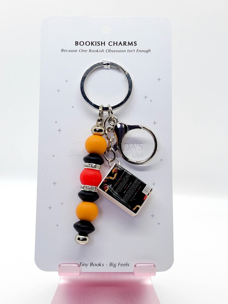 May include: A keychain with a silver ring and a charm featuring a miniature book and a beaded accent. The beads are black, orange, and red, with silver accents. The card reads "BOOKISH CHARMS" and "Tiny Books - Big Feels."