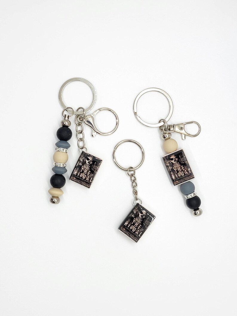 May include: Three keychains with silver-tone rings and clasps. Each keychain features a small, rectangular charm with the text "THE RITUAL" and a skull image. Two keychains have beaded accents in black, gray, and cream colors.