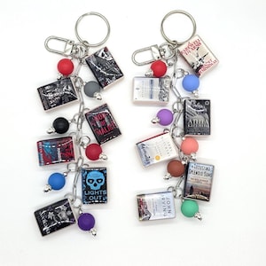 May include: Two keychains, each with miniature book charms. The charms feature book titles like "Lights Out" and "Anna Karenina," along with colorful beads and silver chain links. The keychains are on a white background.