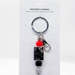 May include: A keychain with a silver ring and clasp, featuring a red bead, a black and white book-shaped charm, a black bead, a silver bead, and a gray bead. The book charm reads "Haunting Adeline". The card reads "Bookish Charms" and "Because One Bookish Obsession Isn't Enough."