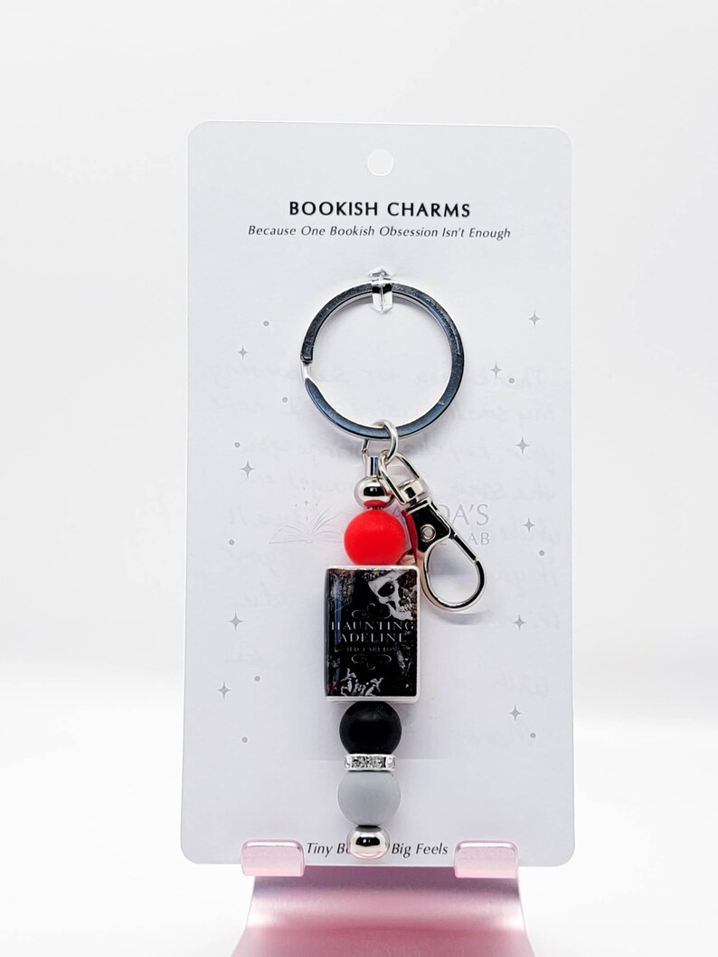 May include: A keychain with a silver ring and clasp, featuring a red bead, a black and white book-shaped charm, a black bead, a silver bead, and a gray bead. The book charm reads "Haunting Adeline". The card reads "Bookish Charms" and "Because One Bookish Obsession Isn't Enough."