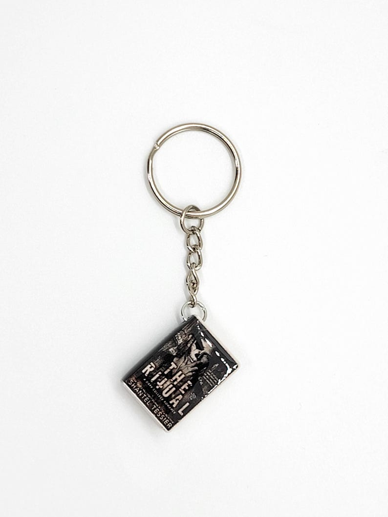 May include: A silver keychain with a rectangular charm featuring the book cover of "The Ritual" by Shantel Tessier. The charm has a black and white design with the book title and author's name visible.