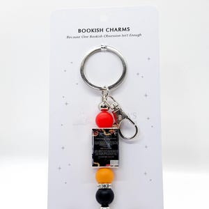 May include: A keychain with a silver ring and clasp, featuring a miniature book charm and colorful beads. The charm includes a small book, red, orange, yellow, and black beads. The card reads "BOOKISH CHARMS" and "Tiny Books - Big Feels".