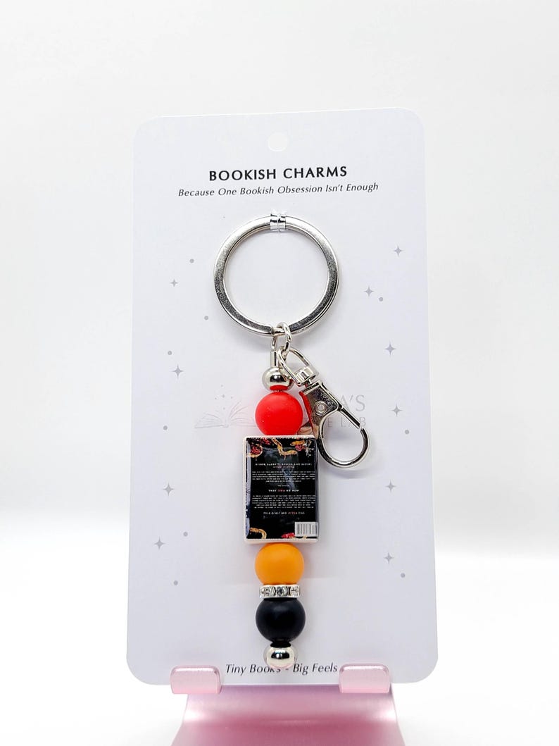 May include: A keychain with a silver ring and clasp, featuring a miniature book charm and colorful beads. The charm includes a small book, red, orange, yellow, and black beads. The card reads "BOOKISH CHARMS" and "Tiny Books - Big Feels".