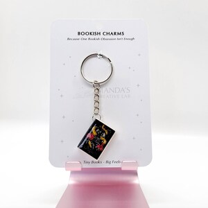 May include: A keychain featuring a miniature book charm with a black background and gold floral accents. The charm hangs from a silver chain and keyring. The card reads "BOOKISH CHARMS" and "Tiny Books - Big Feels."