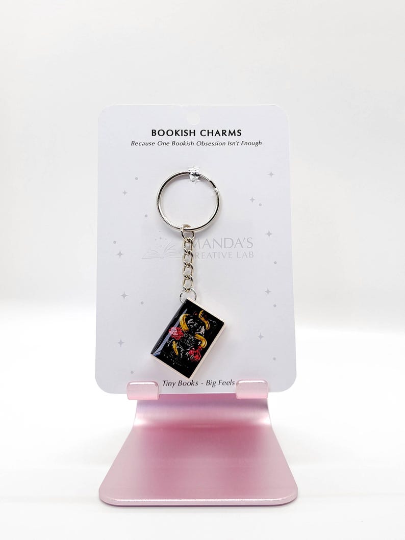 May include: A keychain featuring a miniature book charm with a black background and gold floral accents. The charm hangs from a silver chain and keyring. The card reads "BOOKISH CHARMS" and "Tiny Books - Big Feels."