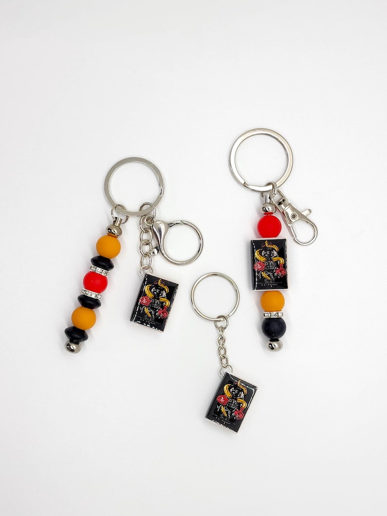 May include: Keychains with black rectangular charms featuring a gold design and red roses. The keychains have silver rings and beaded accents in black, red, and yellow. One keychain has a lobster clasp. The background is plain white.