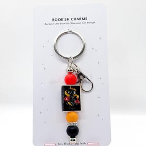 May include: A keychain with a silver ring and clasp, featuring a small book charm with a snake and rose design. The keychain also includes red, orange, black, and silver beads. The text on the card reads "BOOKISH CHARMS" and "Tiny Books - dig Feels."