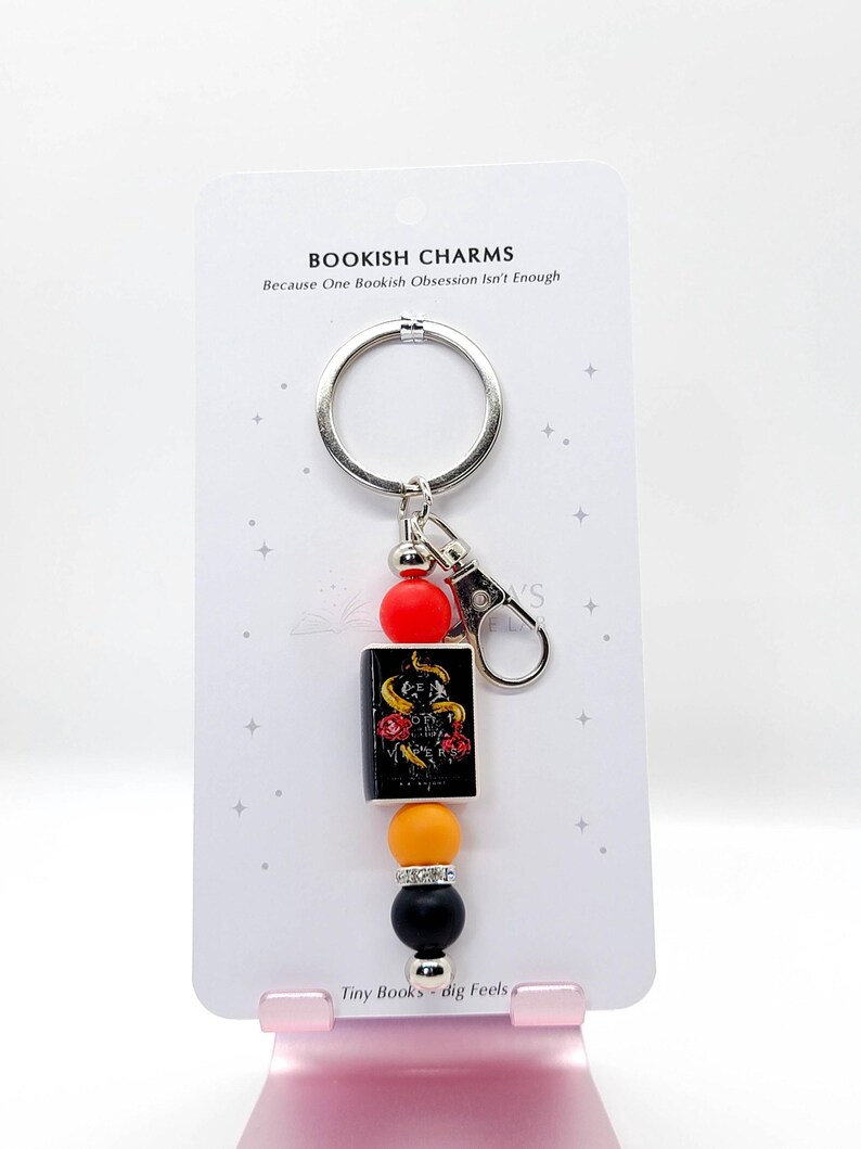 May include: A keychain with a silver ring and clasp, featuring a small book charm with a snake and rose design. The keychain also includes red, orange, black, and silver beads. The text on the card reads "BOOKISH CHARMS" and "Tiny Books - dig Feels."