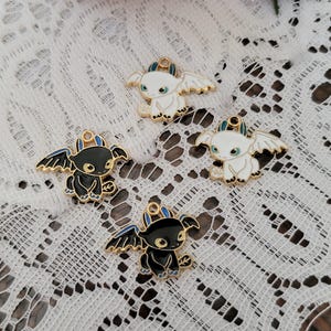 May include: Four enamel dragon charms in black and white with gold detailing. The charms have detailed wings and a loop for attaching to jewellery or crafts. The background is a white lace fabric with a pink rose.