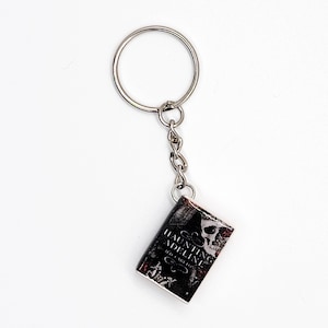 May include: A silver keychain with a small, rectangular charm. The charm is a miniature book with the title "Haunting Adeline" in white text, a skull graphic, and a dark, textured background. The keychain is on a white background.