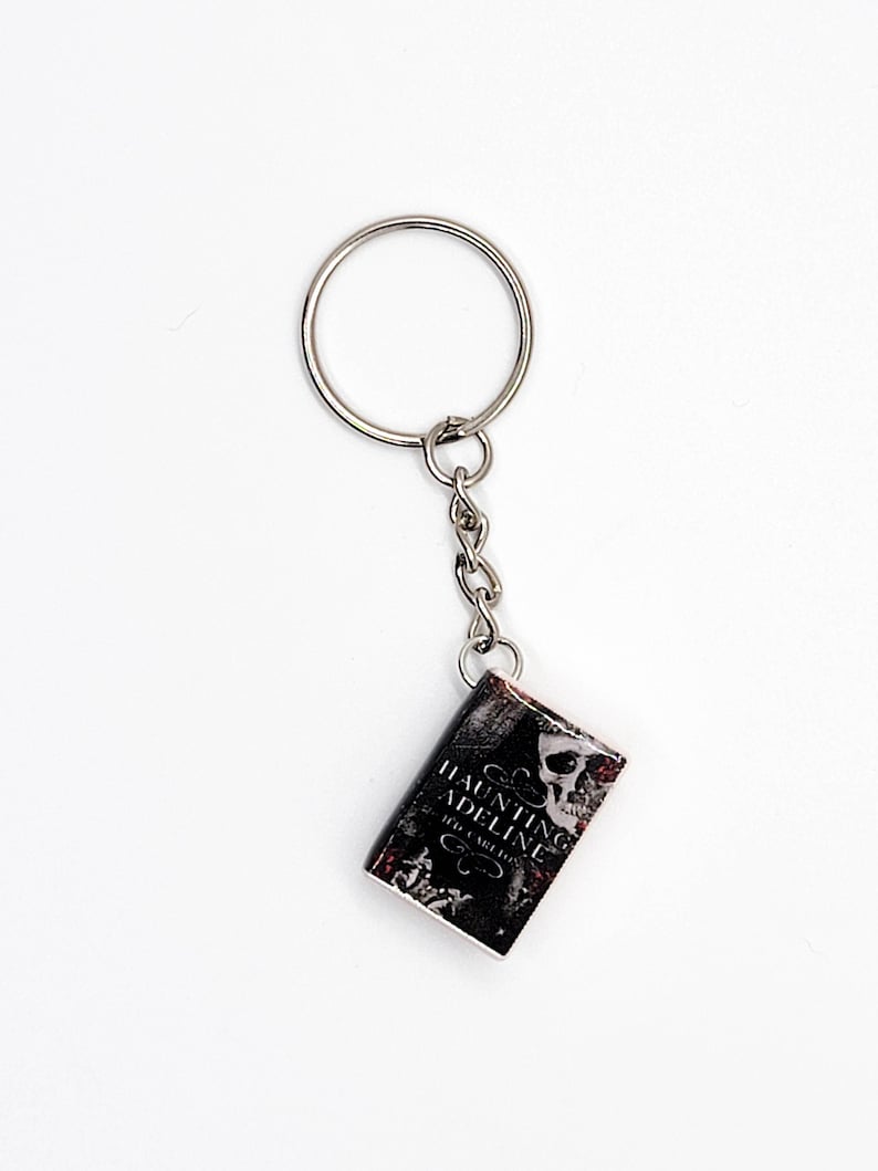 May include: A silver keychain with a small, rectangular charm. The charm is a miniature book with the title "Haunting Adeline" in white text, a skull graphic, and a dark, textured background. The keychain is on a white background.