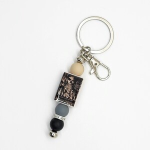 May include: A silver keychain with a lobster clasp and a charm featuring a miniature book titled "THE RITUAL". The charm is accented with a beige, gray, black, and silver bead design.