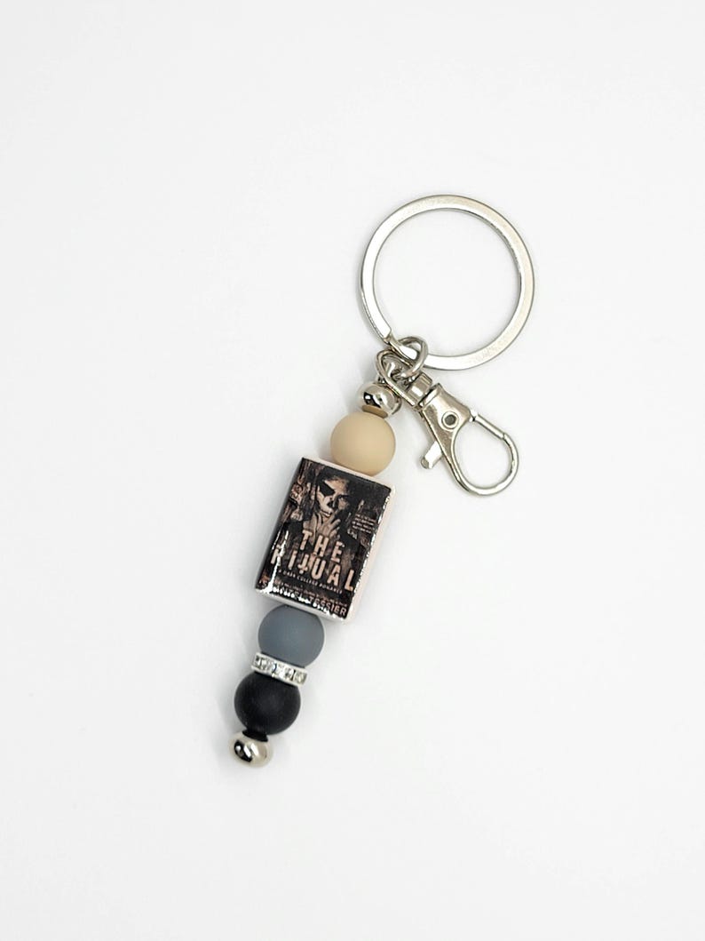 May include: A silver keychain with a lobster clasp and a charm featuring a miniature book titled "THE RITUAL". The charm is accented with a beige, gray, black, and silver bead design.
