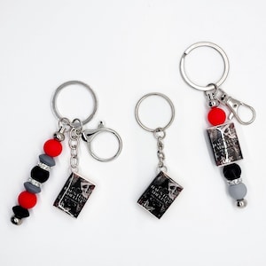 May include: Three keychains with silver rings and lobster clasps. Each keychain features a small book charm with the text "Haunting Adeline" and a skull graphic. One keychain has red, black, and gray beads.