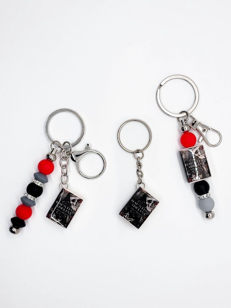 May include: Three keychains with silver rings and lobster clasps. Each keychain features a small book charm with the text "Haunting Adeline" and a skull graphic. One keychain has red, black, and gray beads.