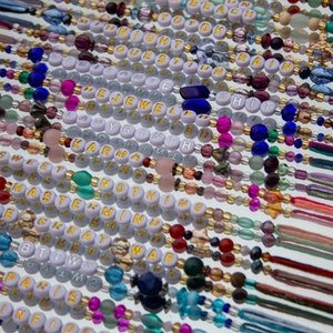 May include: A close-up of a collection of colorful beaded bracelets with gold lettering spelling out words like "karma", "mastermind", "nothing", and "the great war".