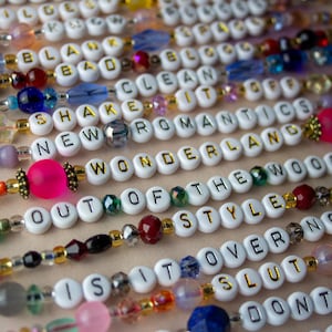 May include: A close-up of colourful beaded necklaces with white letter beads spelling out phrases like "New Romantics", "Wonderland", "Out of the Woods", and "Is it Over Now".