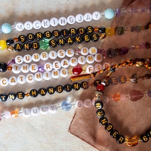 May include: A close-up of six beaded bracelets with different colored beads and letter beads spelling out phrases from the Hunger Games book series. The phrases are "Mockingjay", "Songbirds and Snakes", "Sunrise on the Reaping", "Catching Fire", "The Hanging Tree", and "Hunger Games".