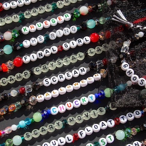 May include: A collection of colorful beaded bracelets with white letter beads spelling out horror movie titles, including "Scream", "Halloween", "Final Girl", "The Ring", and "Ghostface".
