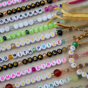 Puede incluir: Una colección de pulseras de cuentas coloridas con palabras como "Dark Academia", "Read More", "Banned Book Club" y "Romance Girlie".