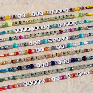 May include: Ten colorful friendship bracelets with different words and beads. The bracelets are made with string and beads, including clear, turquoise, red, orange, yellow, green, and blue beads. The words on the bracelets include "NO ITS", "KEY LIME GREEN", "SEEMINGLY RANCH", "SHIMMER", "YES WHALE", "87 89", "MY MIND IS ALIVE", "123 LGB", "INCANDESCENT GLOW", and "KARMA IS A CAT".