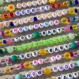 Könnte beinhalten: Eine Sammlung bunter Perlenarmbänder mit Buchstabenperlen, die Sätze wie "Groovy", "Book It", "Be Kind Rewind", "Nostalgia", "80s Baby", "Goosebumps", "90s Baby", "As If", "How Rude" und "Baby" buchstabieren.