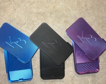 May include: Three phone cases in blue, black, and purple. Each case has a matching lid with the embossed letters "KB". The cases are made of a ridged material and are open, revealing the interior.