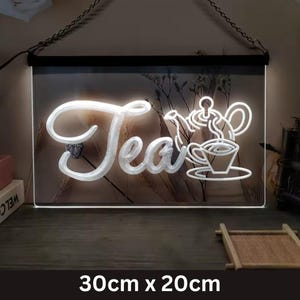May include: A rectangular LED sign with the word "Tea" in a cursive font, accompanied by a teapot and teacup design. The sign is 30cm x 20cm and hangs from a chain. The sign is white and has a black border.