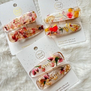 May include: Set of three clear hair clips with pressed flowers and gold lettering. The clips are labeled "May", "Nonny", and "Katie".