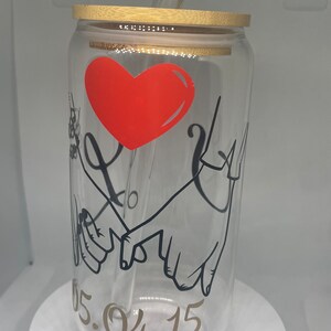 Peut inclure: Un gobelet transparent en forme de canette avec un couvercle en bambou et une paille transparente. Le gobelet présente un cœur rouge et un dessin au trait noir de mains qui se tiennent. La date "05.04.15" est imprimée en or.