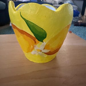 May include: A hand-painted yellow ceramic planter with a lemon design. The planter features a bright yellow base with a lemon slice design, a green leaf, and a white flower. The planter is sitting on a wooden surface.