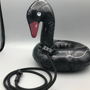 May include: Black inflatable swan pool float with a red beak and white eye details. The float has a black rope attached with a black plastic clasp. The swan has white wave-like accents around the float.