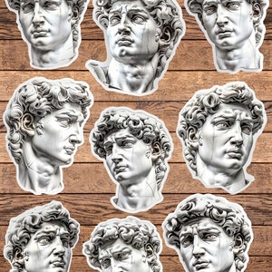 May include: A collection of white stickers featuring the sculpted head of David, a Renaissance masterpiece. The stickers showcase the detailed facial features and curly hair of the statue, set against a wooden background. Ideal for art enthusiasts and sticker collectors.
