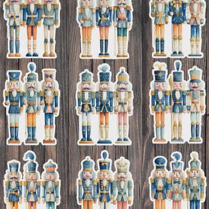 May include: A collection of watercolor-style nutcracker soldier stickers. Each sticker features a different design with varying uniforms in shades of blue, gold, and red. The stickers are arranged on a wooden background.
