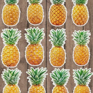 May include: A collection of pineapple stickers. Each sticker features a detailed illustration of a ripe pineapple with a textured, golden-yellow body and a crown of green leaves. The stickers are set against a wooden background.